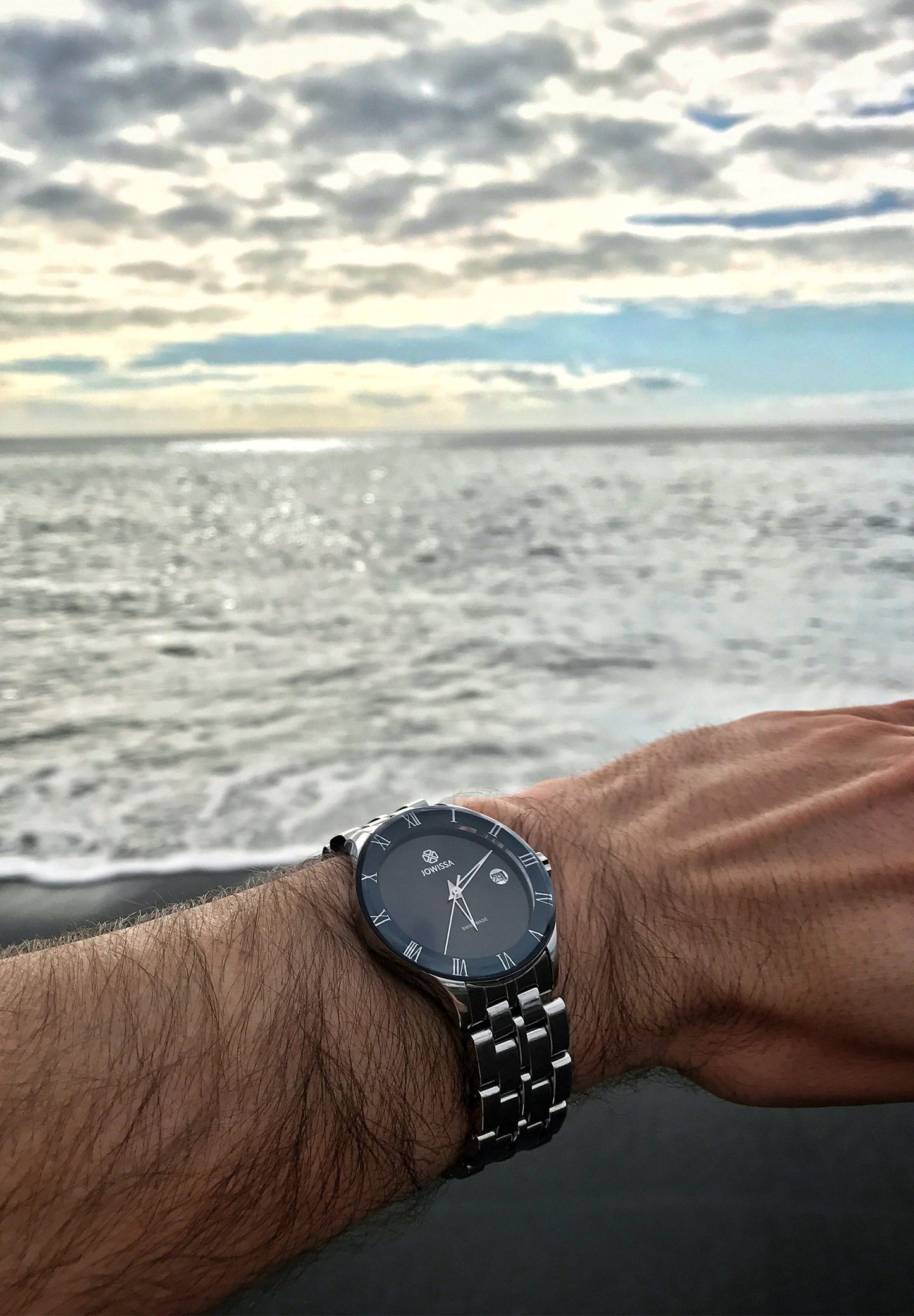 Silver swiss made watch worn by a man on the beach - The shiny sunray dial, along with filigree Roman numerals indices and triangular, hollow Dauphine-style hands, adds a touch of sophistication. The tempered mineral glass with a polished edge provides robust protection, while its 5 ATM water resistance ensures it's suitable for everyday activities, including showers.