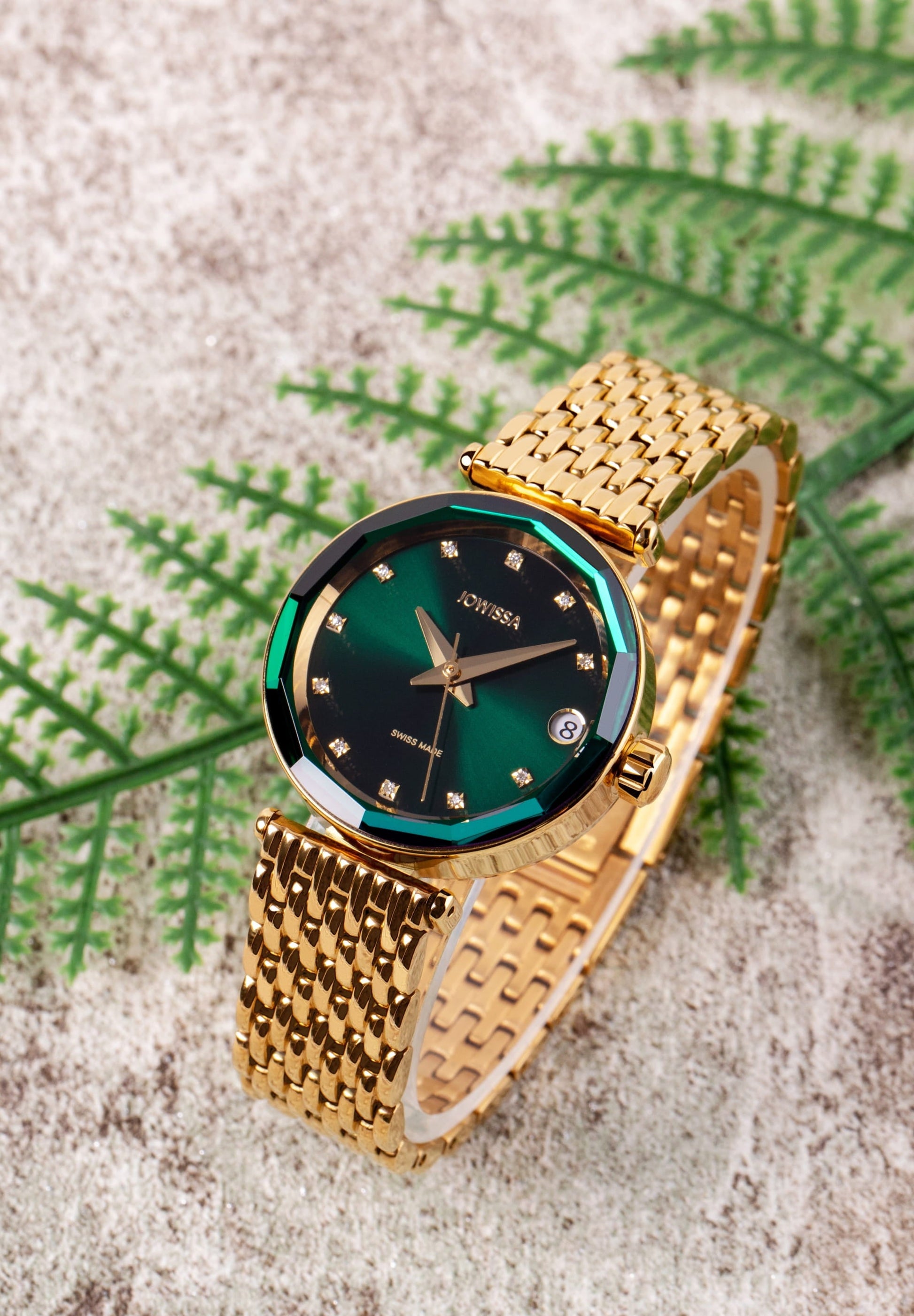 Swiss Automatic womens Watch- a luxurious women's timepiece. With a stainless steel case plated in gold and green sunray dial - It has11 diamond hour markers and a scratch-resistant sapphire crystal with a diamond-cut bezel for added sparkle. The watch is placed on the sand with a green leaf