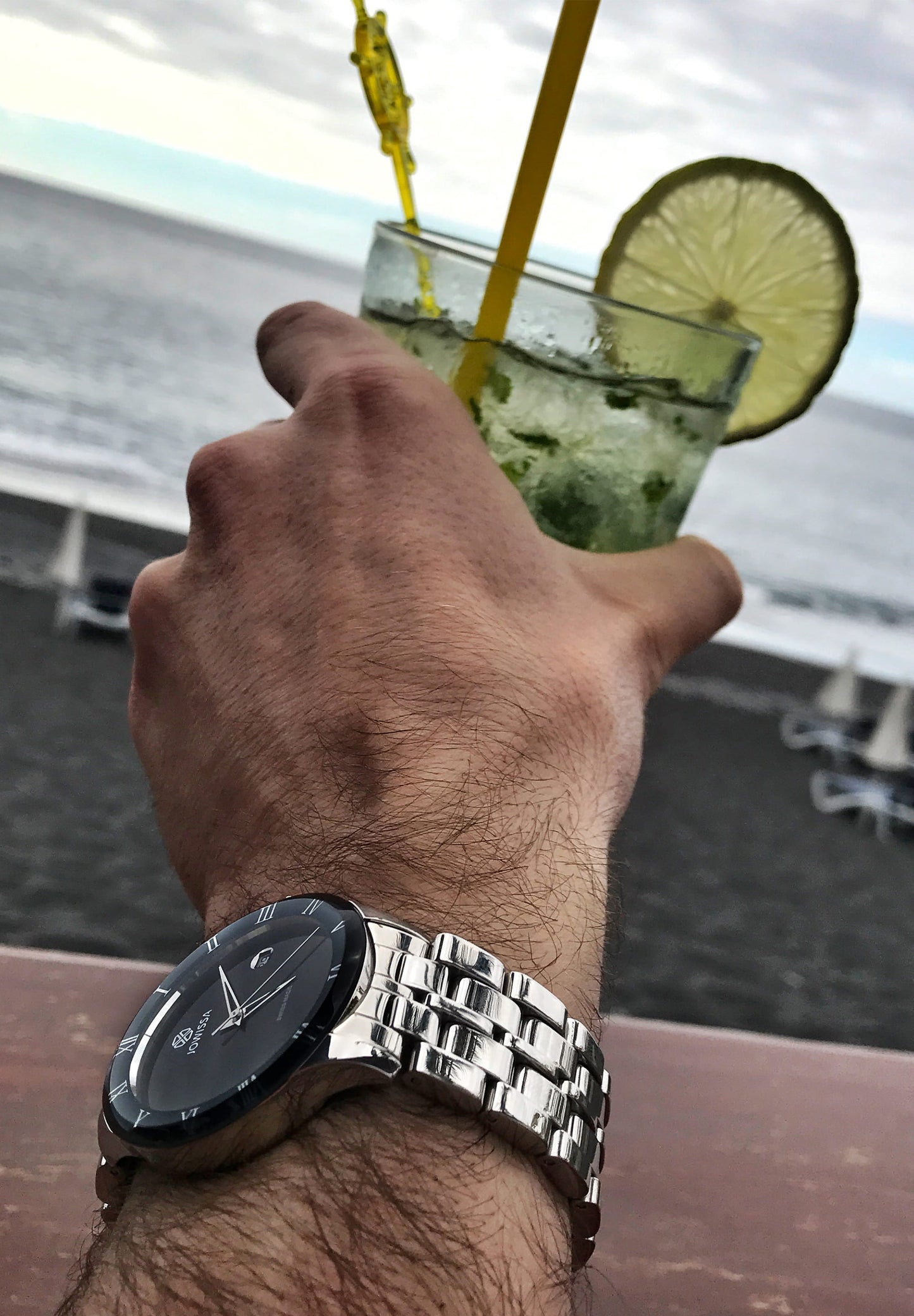 Silver watch with black details worn by a man holding a cocktail on the hand on the beach. Swiss-made watch features a polished stainless steel case and bracelet, making it both durable and stylish. All swiss made