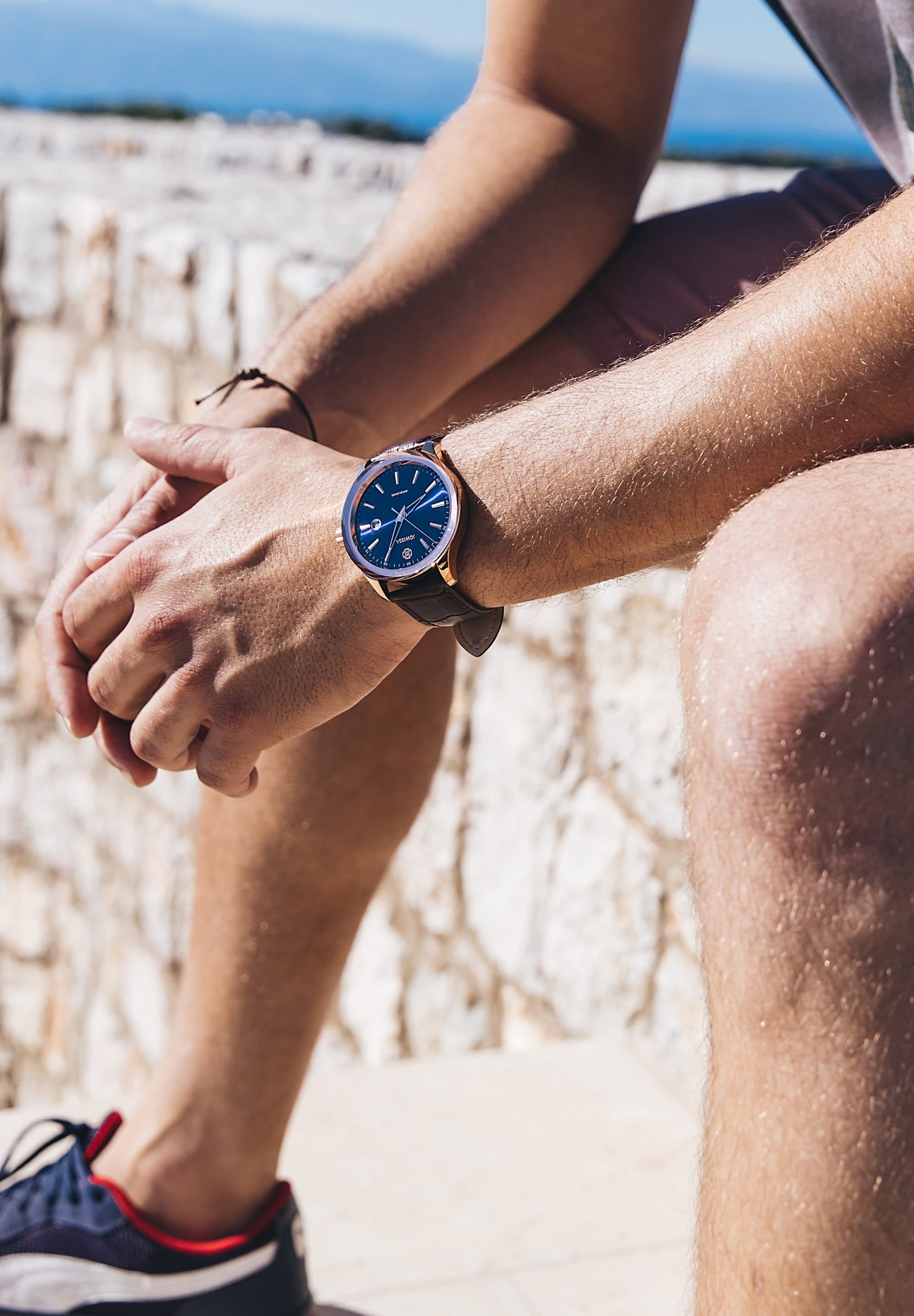 A man sitting comfortably, showcasing the Tiro Swiss Men's Watch J4.203.L on his wrist, featuring a striking Atlantic blue sunray dial and brown leather strap - all swiss made